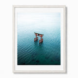 Framed wall art featuring an aerial photograph of a red torii gate in Japan standing in calm blue-green water, presented in a white frame with a white mat against a light gray background.