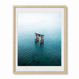 Framed wall art featuring an aerial photograph of a red torii gate in Japan standing in calm blue-green water, presented in a light wood frame with a white mat against a light gray background.