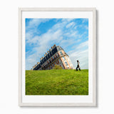A framed art print featuring the sinking of house of Montmartre in Parisg on a grassy hill under a blue sky with white clouds, with a person walking on the grass. The print is framed in a white frame with a white mat against a white background.