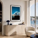 A framed vertical art print of the Sydney Harbour Bridge at night hangs on a beige wall in a modern, sunlit living room. The print features a silhouette of a person standing on the bridge against a deep blue sky and water.