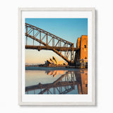 Framed photographic art print of the Sydney Harbour Bridge and Opera House at sunset, shown in a white frame against a light gray background.