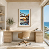 A framed art print of the Sydney Opera House and Harbour Bridge at sunset, displayed on a beige wall above a light wood desk and a woven office chair in a bright, modern home office.