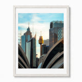 A framed art print of the Sydney skyline, featuring the Sydney Opera House, Sydney Tower, and skyscrapers under a blue sky, presented in a white frame with a white mat.