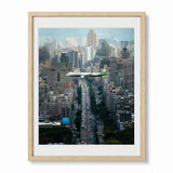 A framed photographic print of a busy city street with an airplane flying over the Taiwan skyline, shown in a light wood frame.
