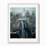 A framed photographic print of a busy city street with an airplane flying over the Taiwan skyline, shown in a white wood frame.