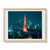 A framed photographic print of the Tokyo Tower at night, illuminated in bright orange-red against a dark blue cityscape, shown in a light natural wood frame with a white mat on a white background.