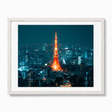 A framed photographic print of the Tokyo Tower at night, glowing orange against a dark blue cityscape, set in a white frame with a white mat.