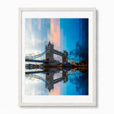 A framed timelapse art print of London's Tower Bridge, featuring a modern overlay of the bridge and sky in shades of blue, orange, and gray, with a reflection in the water. The print is framed in a white frame with a white mat, set against a plain white background.