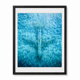 Framed wall art featuring an aerial photograph of an abandoned airplane seen from above, with rippling water patterns across the print. The artwork is presented with a white mat and a black frame against a clean white background.