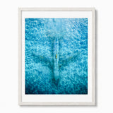 Framed wall art featuring an aerial photograph of an abandoned airplane seen from above, with rippling water patterns across the print. The artwork is presented with a white mat and a white frame against a clean white background.