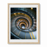 A framed art print featuring a high-angle photograph of the famous bramante staircase in Vatican City, enclosed in a light wood frame