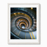 A framed art print featuring a high-angle photograph of the famous bramante staircase in Vatican City, enclosed in a white frame.